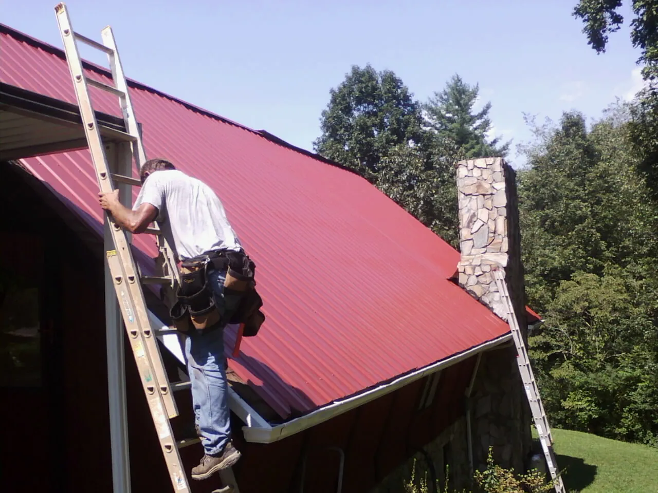 Roofer on a ladder beside a red metal roof and stone chimney — Soffit & Fascia Repair by Roofing Brothers in Lake Forest